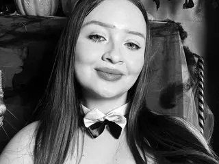 JennaCarsson poses with long dark hair and a slight smile, wearing a white bow neck tie or choker against a dark background.