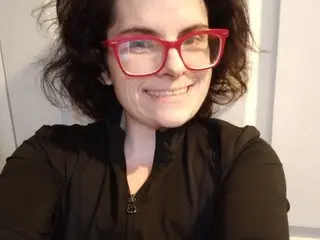 JessaBella smiles at the camera, wearing red-framed glasses and a black zip-up jacket with her dark hair styled up.