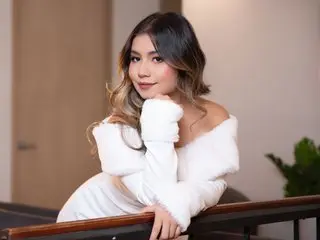 JessieOlivetti poses with her hands under her chin, wearing a white off-the-shoulder top and letting her wavy brown hair fall over her shoulders.