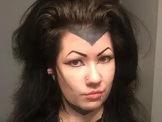 Woman with Maleficent cosplay makeup, dark wavy hair, and dramatic widow's peak facial tattoos.