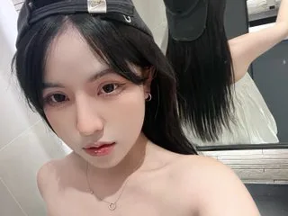 JoiceBae looks directly at the camera with long black hair and red lipstick, showing her bare shoulders and neck.
