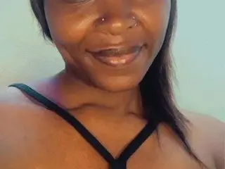 Smiling Black woman in a black tank top posing for a selfie against a light green wall.