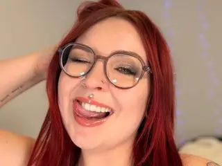 Smiling woman with red hair and glasses laughing in a cozy indoor setting.