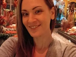 JoyCherrystone smiles at the camera, showing her face and long brown hair while wearing a grey knit top.