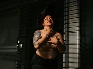 JuanaValkiria poses with her eyes closed, wearing a tan tank top and showing off her arm tattoos while holding her hands near her chest.