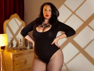 JudithConner poses in a black bodysuit and black fishnet tights, showing her cleavage and dark hair against a warm, indoor background.