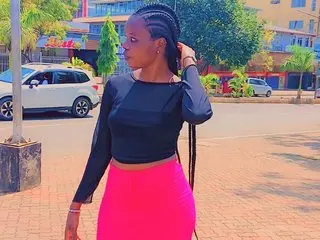 JuicyAssBarbie poses outdoors wearing a black long-sleeved top and bright pink skirt, showing off her long dark braided hair.