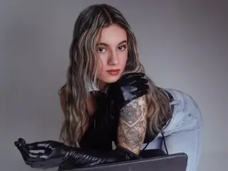 JuliaColeman poses with long wavy hair, wearing black gloves and a black top, looking directly at the camera against a plain grey background.