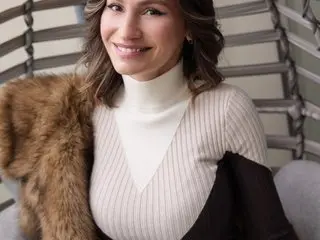 JuliaRichard smiles while wearing a white turtleneck and a brown fur coat, showing her wavy brown hair and shoulders.