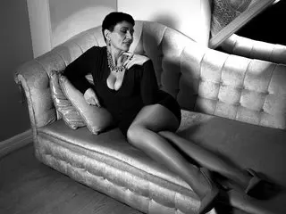 JuliaWinslet poses naked on a light-colored tufted sofa, wearing a black top that shows her cleavage, with her legs extended and hair pulled back.