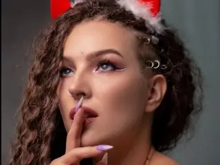JulietaLane poses with long, curly brown hair and winged eyeliner, looking off-camera with a hand near her lips.