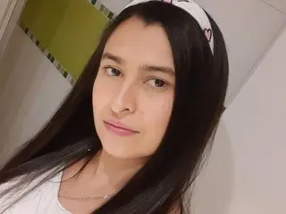 JulietaRodriguez poses with her long dark hair down, wearing a white top against a plain wall background.