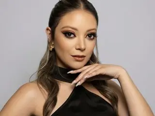 JulietaSadovnik poses with a hand under her chin, wearing a black halter top and showing her long, wavy brown hair against a plain grey background.