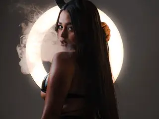 Juliii_Sweet poses with long black hair and a black bra, looking over her shoulder against a bright circular light.