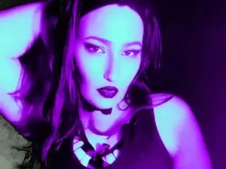 JunoJade poses with her hands behind her head, wearing a black top and dark lipstick against a dark background.