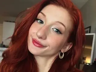 JustALilBrat, a redheaded woman, smiles at the camera with a close-up view of her face and long red hair.