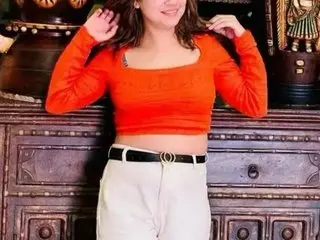 JyotiiAngel poses wearing a tight orange long-sleeve top and white pants, running her hands through her long dark hair against a dark wooden cabinet.