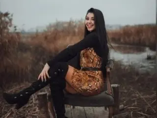 KIYAARA smiles while sitting on a wooden bench outdoors in a field, wearing a black long-sleeve top, a patterned mini dress, and tall black boots.