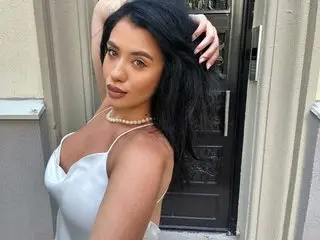 Kaithvonteasee poses with long black hair, wearing a white tank top that shows her shoulders and cleavage against a neutral wall background.