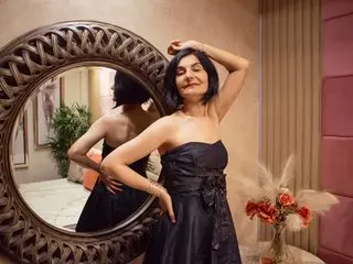 KalystaSwiss poses with one hand behind her head, wearing a black strapless dress against a round mirror and warm indoor background.