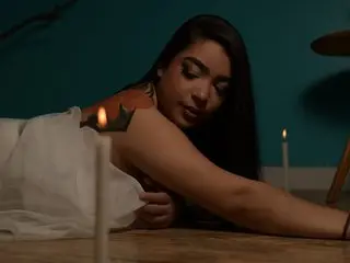 KamilaBetancourt poses with long dark hair, leaning forward over a surface near a lit candle in a dim setting.