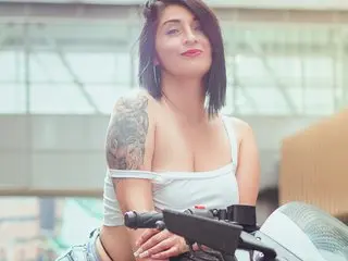 Karen_lennox poses with her dark hair down, wearing a white tank top that shows her shoulder and chest, with a large tattoo visible on her arm.