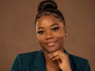 KarinaMillers poses with a soft smile, wearing a dark teal blazer and her hair styled in a neat, high bun against a plain brown background.