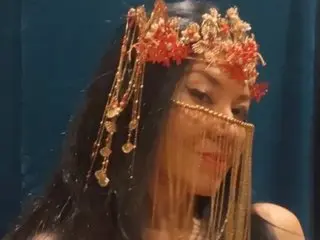 Karma_Queen poses with long dark hair and a gold, beaded headpiece and face jewelry against a dark blue background.