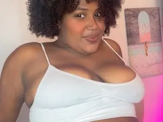 Smiling woman with curly hair wearing a white camisole in a cozy, brightly lit room.