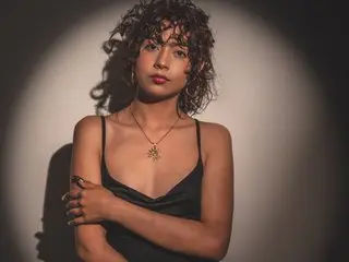 Young woman with curly hair in a black satin camisole posing under dramatic spotlight lighting.