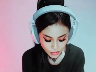 KatherinBess looks down with her eyes closed, wearing large white headphones and a black top against a soft pink and blue background.