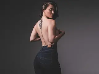 Katt_Henao poses shirtless with her back turned, showing her bare back and shoulder tattoo against a dark background.
