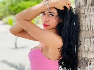 Katte_D poses with her hands behind her head, showing her long black hair and pink top against a bright outdoor background.