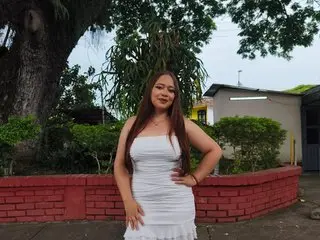 Katty_rose99 poses outdoors in a white strapless dress, showing her bare shoulders and chest against a background of green trees and a house.