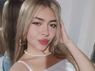 KatyBennet poses with her blonde hair swept back, wearing a white top and a pearl necklace against a plain background.