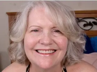 Smiling mature woman with blonde hair in a cozy bedroom setting.