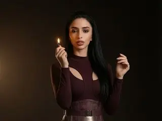 KaylaJoyy poses with long black hair, wearing a dark maroon long-sleeve top against a dark background while holding a lit candle.