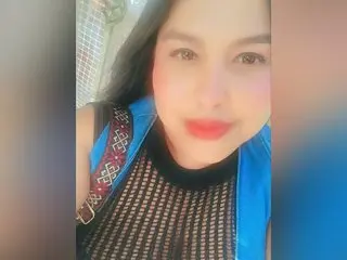 Smiling woman in a black mesh top and blue vest posing for a casual outdoor selfie.