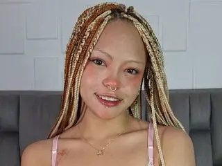 Smiling woman with blonde braids and nose ring wearing a pink camisole in a modern room.