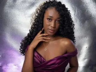 KendallMay poses with her long, dark curly hair down, wearing a purple satin off-the-shoulder top against a soft, light grey background.
