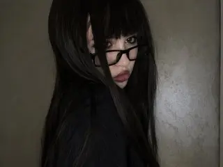 KendyMiller poses with long black hair and black glasses, looking directly at the camera against a plain wall.