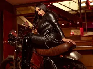 KeyshaTyler poses naked in black leather gear, sitting on a motorcycle with her hair down in a dark, indoor setting.