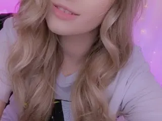 Keziah shows off her long blonde wavy hair while wearing a light purple t-shirt in a soft-lit room.