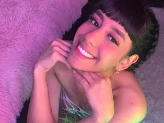 KhloeSmiith smiles while posing with her hands near her face, showing her dark hair and bare shoulders against a soft purple background.