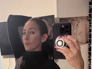 KickassMILF poses with her hands behind her head, wearing a black top against a plain white wall.