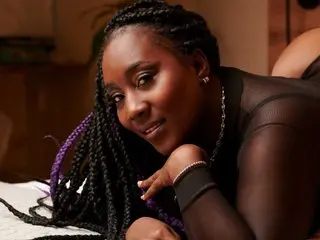 Smiling Black woman with long braids wearing a black sheer top in a cozy indoor setting.