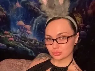 KikkaKink wears glasses and a black choker, posing with her dark hair pulled back against a colorful, trippy mushroom-themed background.