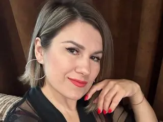 KiraRoxx poses with a smile, wearing a black sheer top, showing her light brown hair and face against a dark brown background.