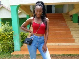 KissMeSlowly22 poses in a red tank top and blue jeans, letting her long dark hair hang down while standing outdoors near a green wall.
