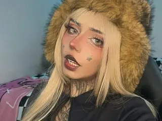 KoraRousee poses with a direct gaze, wearing a blonde wig and a brown furry hood or hat.
